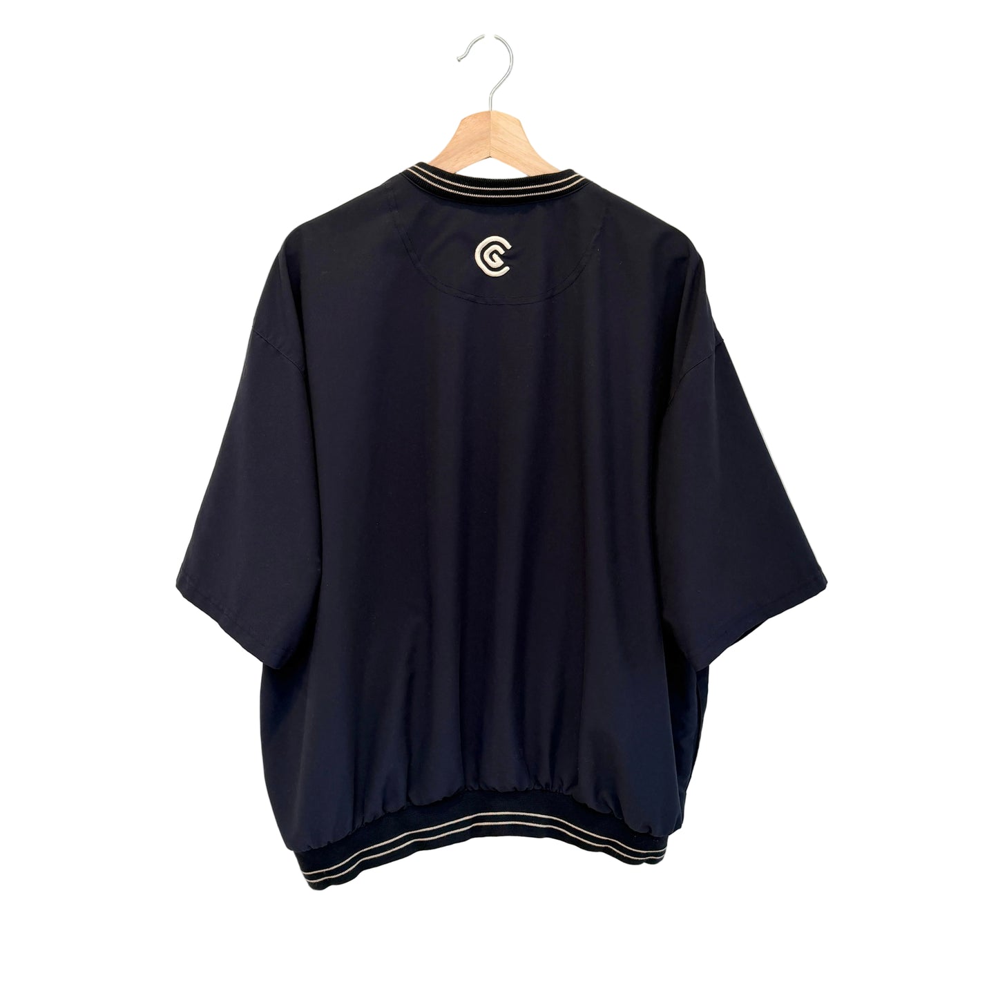 A navy blue short-sleeve bomber jacket with white striped trim on the collar and hem, hanging on a wooden hanger against a white wall. A white circular logo is printed on the upper back.