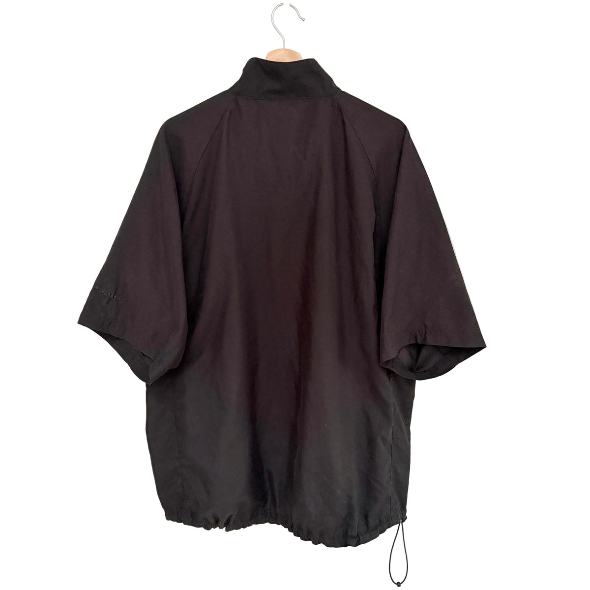 A black short-sleeve high-neck jacket or windbreaker hanging on a wooden hanger against a plain off-white wall.