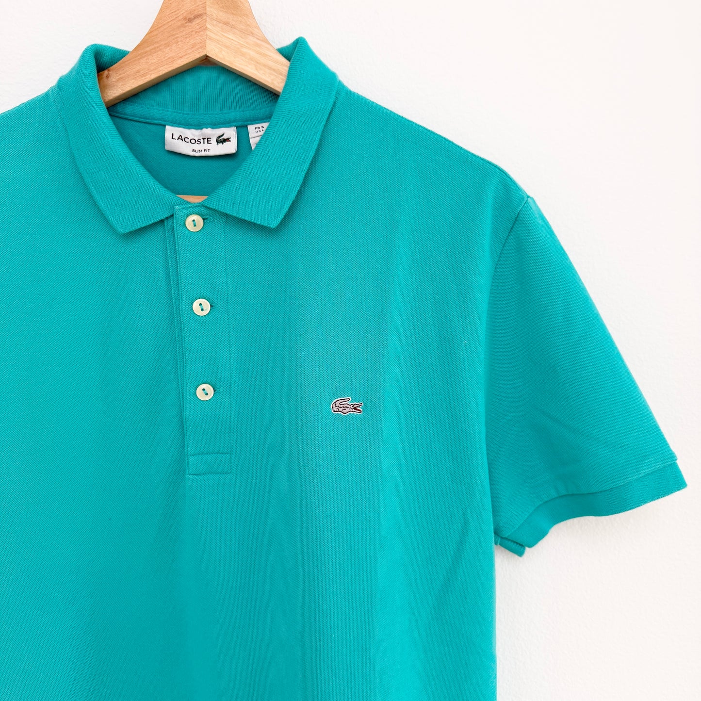 Teal polo shirt with Lacoste logo on a hanger against a white background