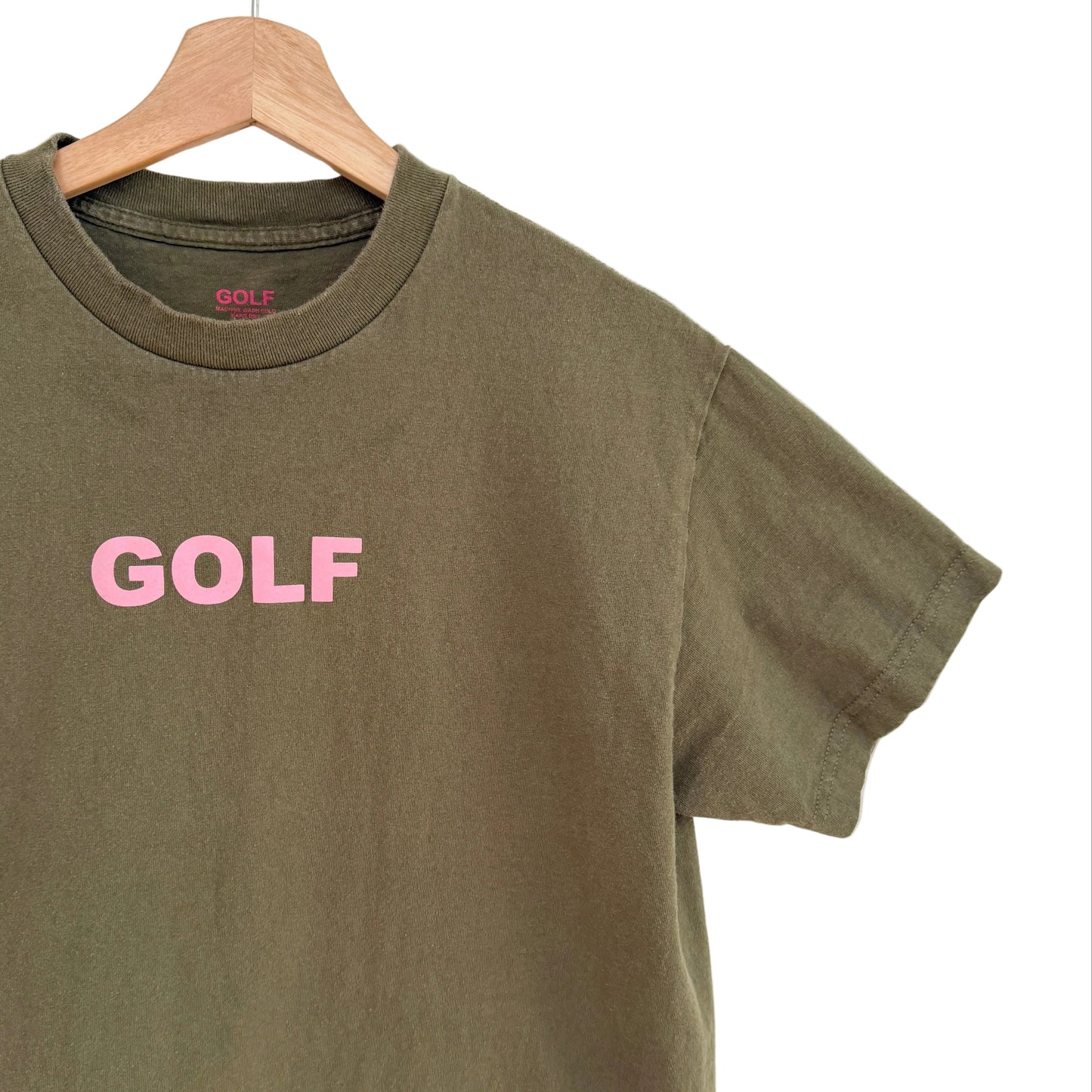Green t-shirt with 'GOLF' printed in pink on a wooden hanger against a white background