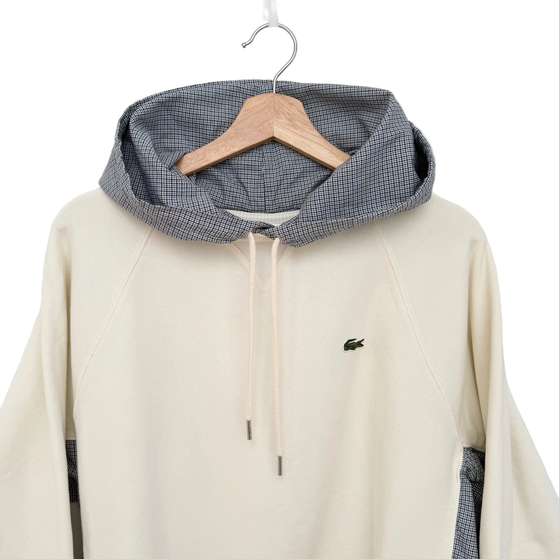 A cream-colored hooded sweatshirt with a gray and white gingham patterned hood lining, hanging on a wooden hanger against a plain white wall. The sweatshirt features a small green Lacoste crocodile logo on the left chest.