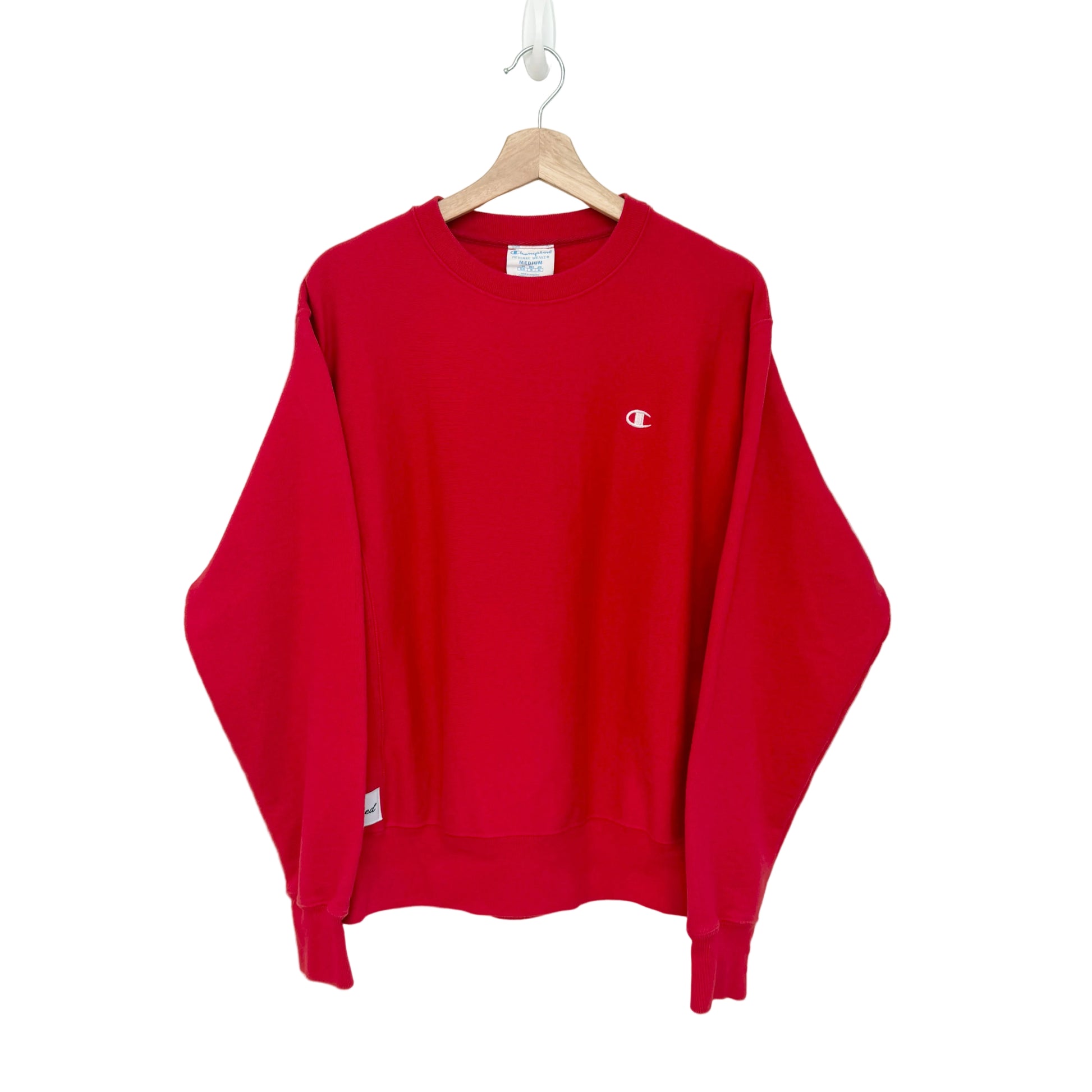 A red Champion crewneck sweatshirt hanging on a wooden hanger against a plain white wall. The iconic small 'C' logo is embroidered on the left chest.