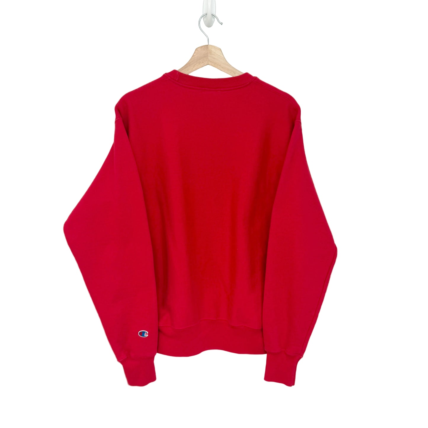 A bright red crewneck sweatshirt hanging on a wooden hanger against a plain white wall. The sweatshirt features the Champion 'C' logo embroidered in blue and red on the left sleeve cuff.