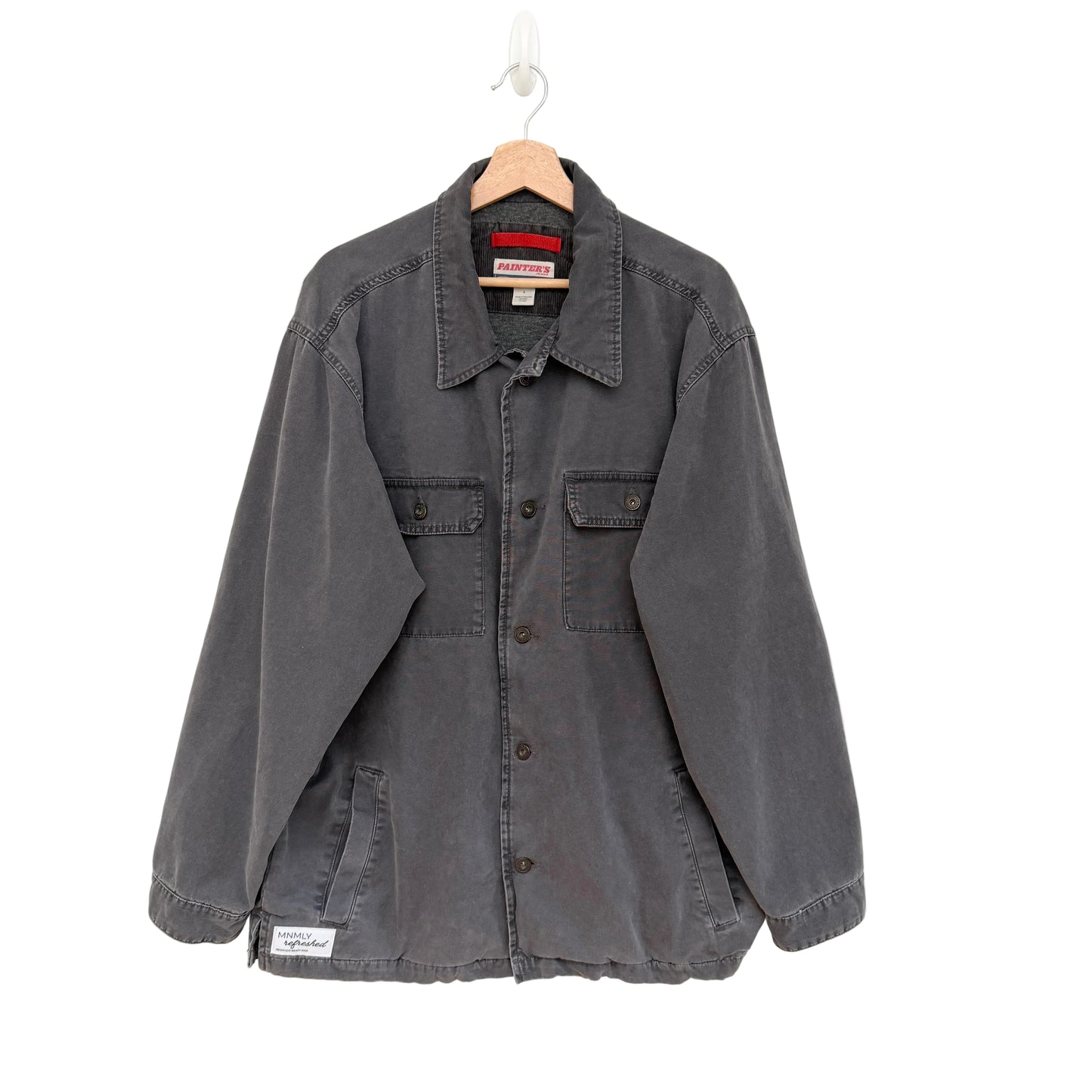 A dark gray washed cotton utility shirt jacket hanging on a wooden hanger against a plain white wall. The jacket features a button-down front, two chest flap pockets, and a small white brand patch on the lower left hem.