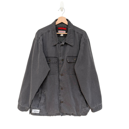 A dark gray washed cotton utility shirt jacket hanging on a wooden hanger against a plain white wall. The jacket features a button-down front, two chest flap pockets, and a small white brand patch on the lower left hem.