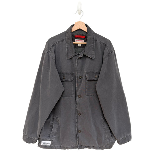 A dark gray washed cotton utility shirt jacket hanging on a wooden hanger against a plain white wall. The jacket features a button-down front, two chest flap pockets, and a small white brand patch on the lower left hem.