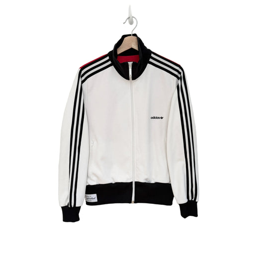 A white Adidas track jacket with black ribbed trim, black three-stripes on the sleeves, and a small black logo on the chest, hanging on a wooden hanger against a plain white wall.