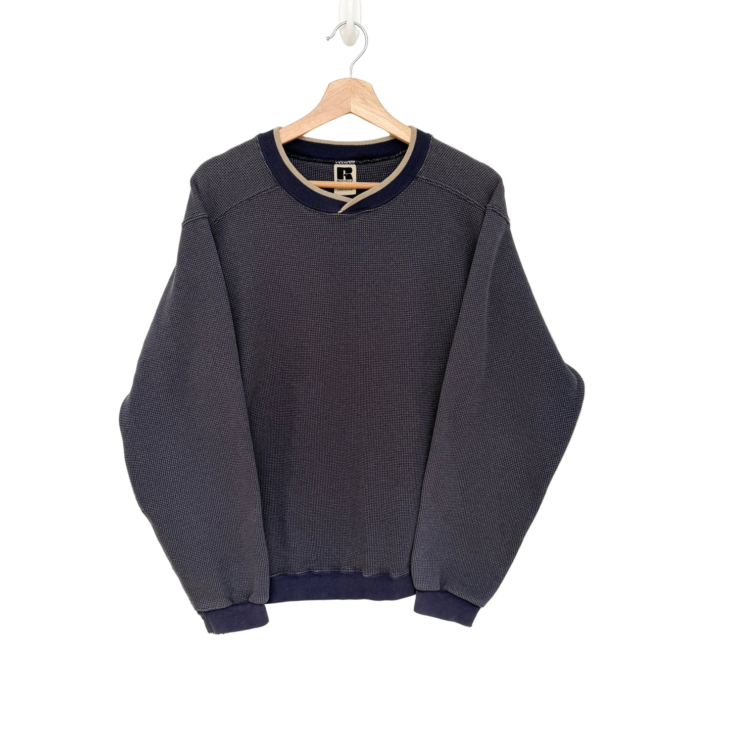 A navy crewneck sweatshirt with navy blue ribbed trim on the collar, cuffs, and hem, hanging on a wooden hanger against a plain white wall.