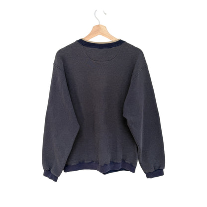A navy crewneck sweatshirt with navy blue ribbed trim on the collar, cuffs, and hem, hanging on a wooden hanger against a plain white wall.