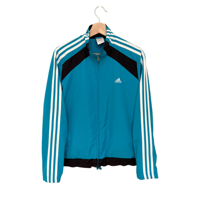 A teal Adidas track jacket with black shoulder panels and white three-stripes on the sleeves, hanging on a wooden hanger against a plain beige wall.