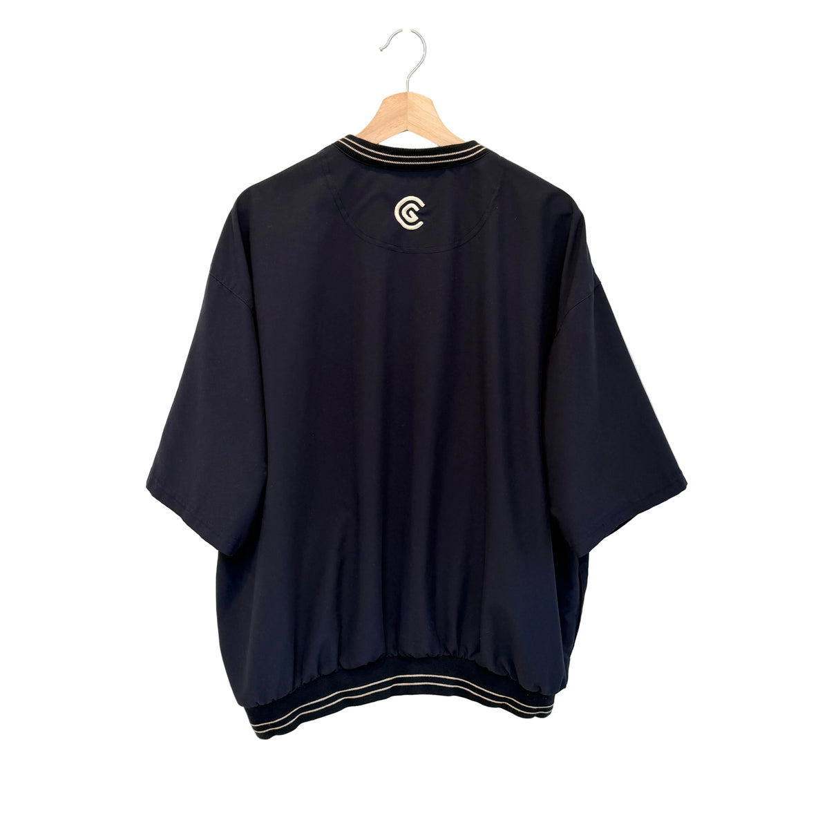 A navy blue short-sleeve bomber jacket with white striped trim on the collar and hem, hanging on a wooden hanger against a white wall. A white circular logo is printed on the upper back.
