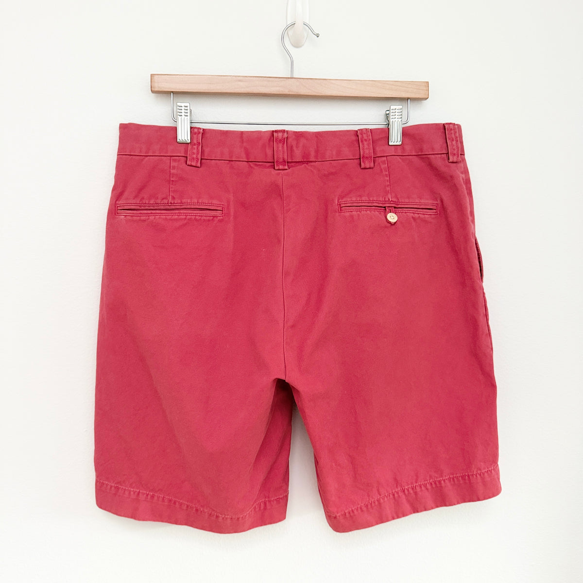 A pair of red chino shorts hanging from a wooden hanger against a plain white wall, showing the back pockets and waistband details.