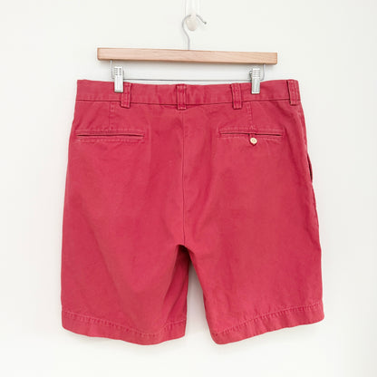 A pair of red chino shorts hanging from a wooden hanger against a plain white wall, showing the back pockets and waistband details.