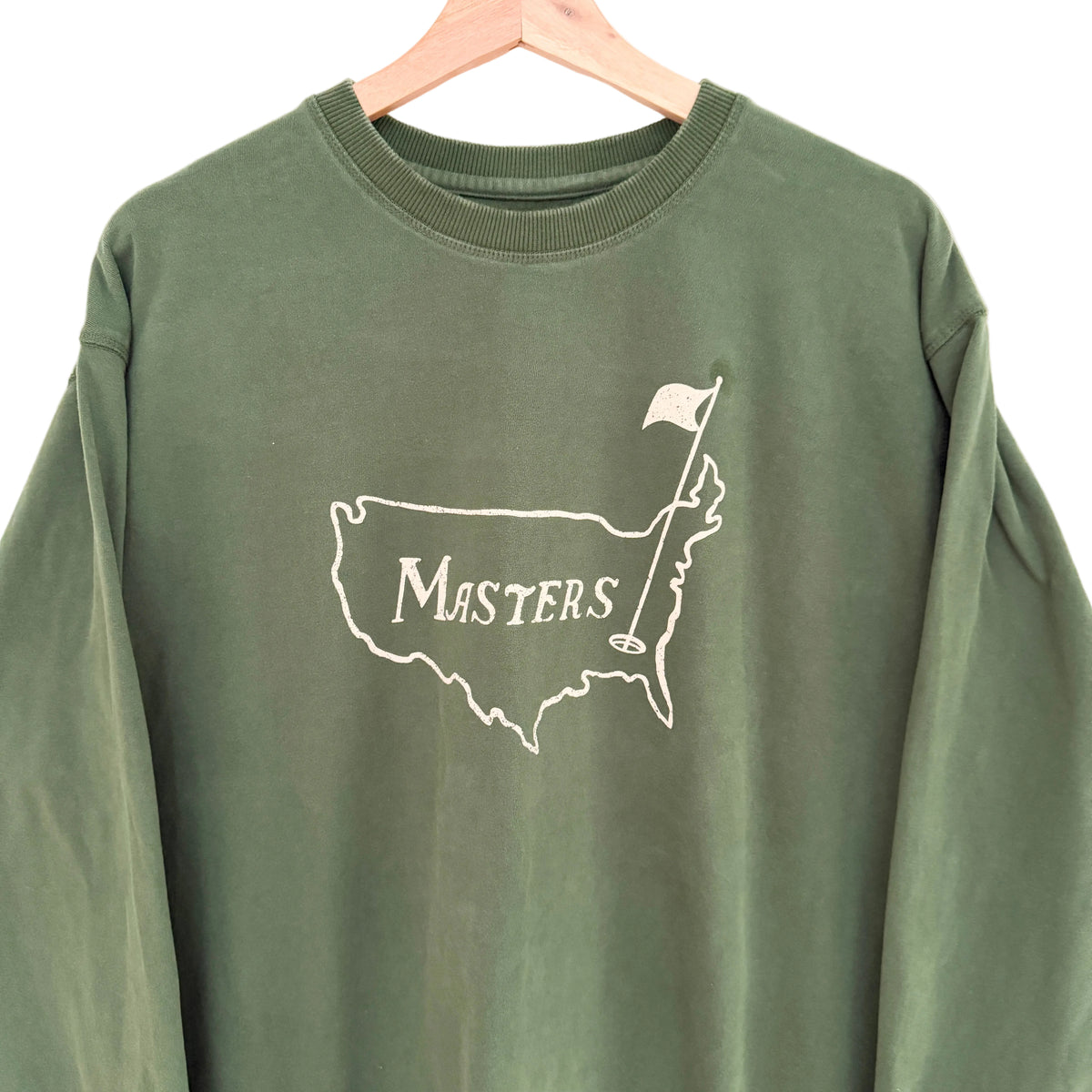 An olive green long-sleeve sweatshirt hanging on a wooden hanger, featuring a white graphic of the Augusta Masters golf course map with a flag and the word 'Masters' on the chest.