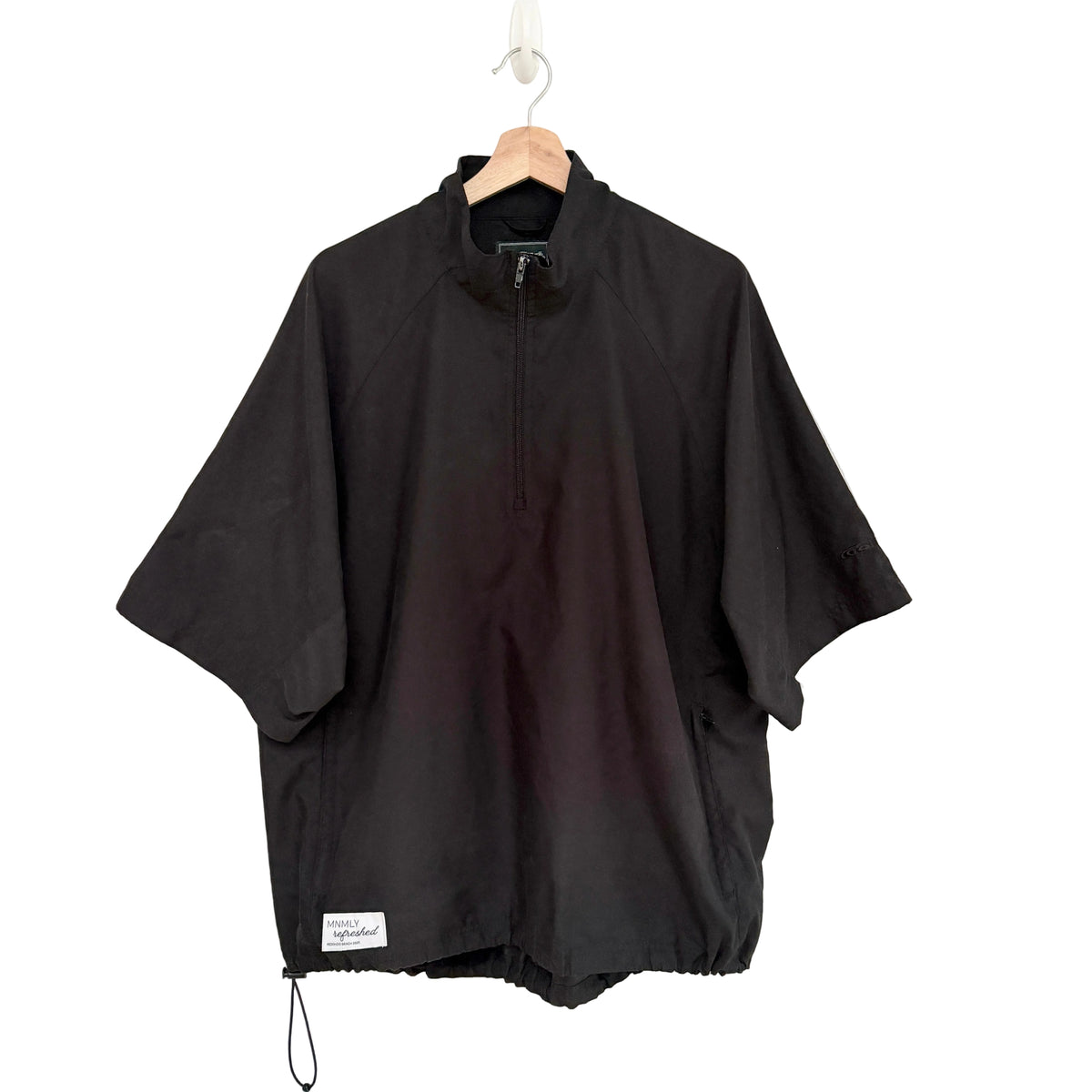 A black half-zip pullover jacket with wide short sleeves, hanging on a wooden hanger against a plain white wall. A small white brand label is visible near the bottom hem.