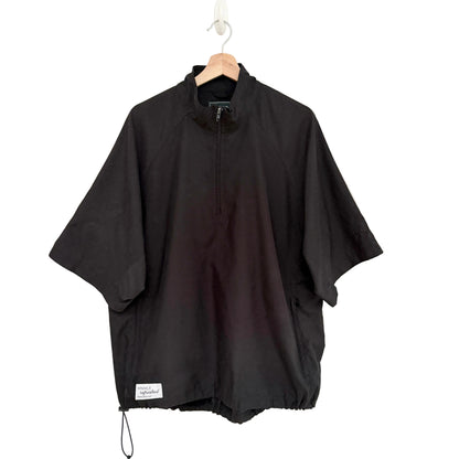 A black half-zip pullover jacket with wide short sleeves, hanging on a wooden hanger against a plain white wall. A small white brand label is visible near the bottom hem.