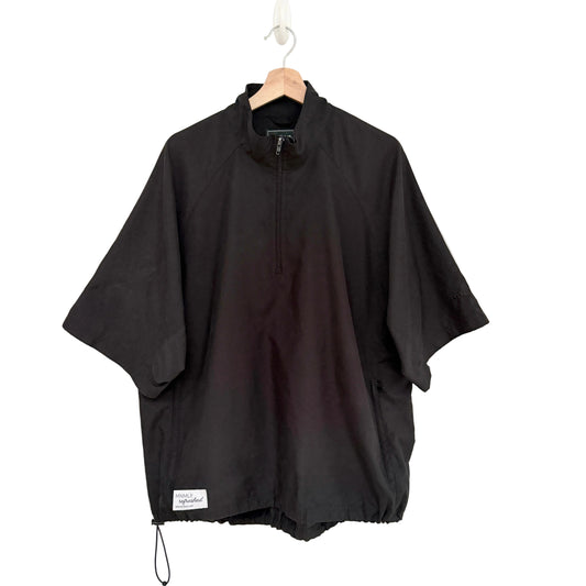 A black half-zip pullover jacket with wide short sleeves, hanging on a wooden hanger against a plain white wall. A small white brand label is visible near the bottom hem.