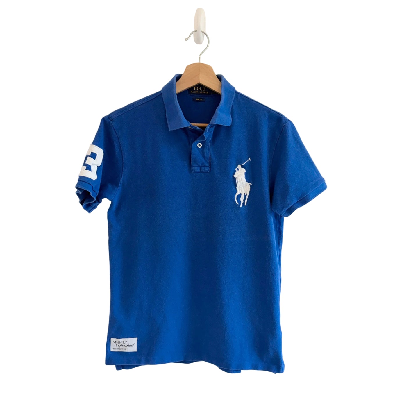 Blue polo shirt with a white logo on a hanger against a beige wall